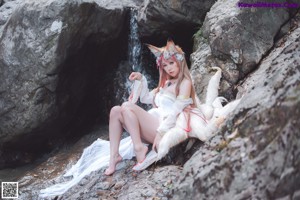A woman in a white dress sitting on a rock next to a stream.